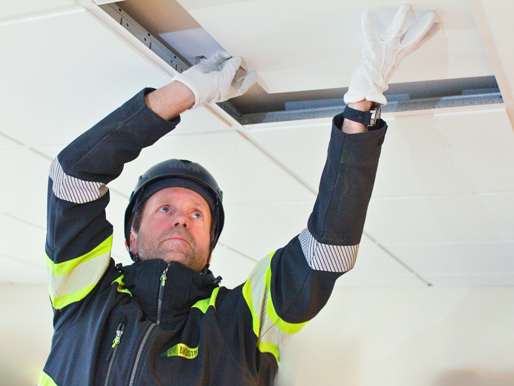 A construction worker wearing a helmet, safety jacket, and white gloves installs or adjusts a ceiling tile in a suspended ceiling grid. The worker is looking up and using both hands to fit the tile into place.